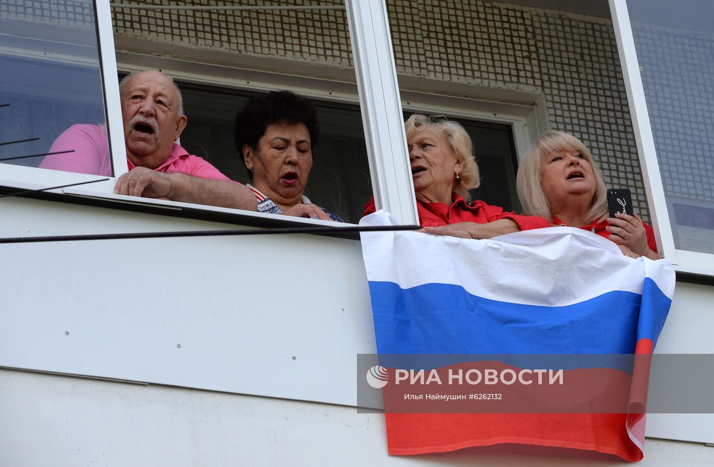 Жители дома на улице Ключевской в Красноярске поют Гимн Российской Федерации вместе с музыкальным ансамблем Главного управления МВД России по Красноярскому краю "Голоса Енисея", который поздравляет горожан, находящихся в своих домах в режиме самоизоляции, с праздником Дня России. Место: Россия, Красноярск. Автор: Илья Наймушин/РИА Новости. Празднование Дня России